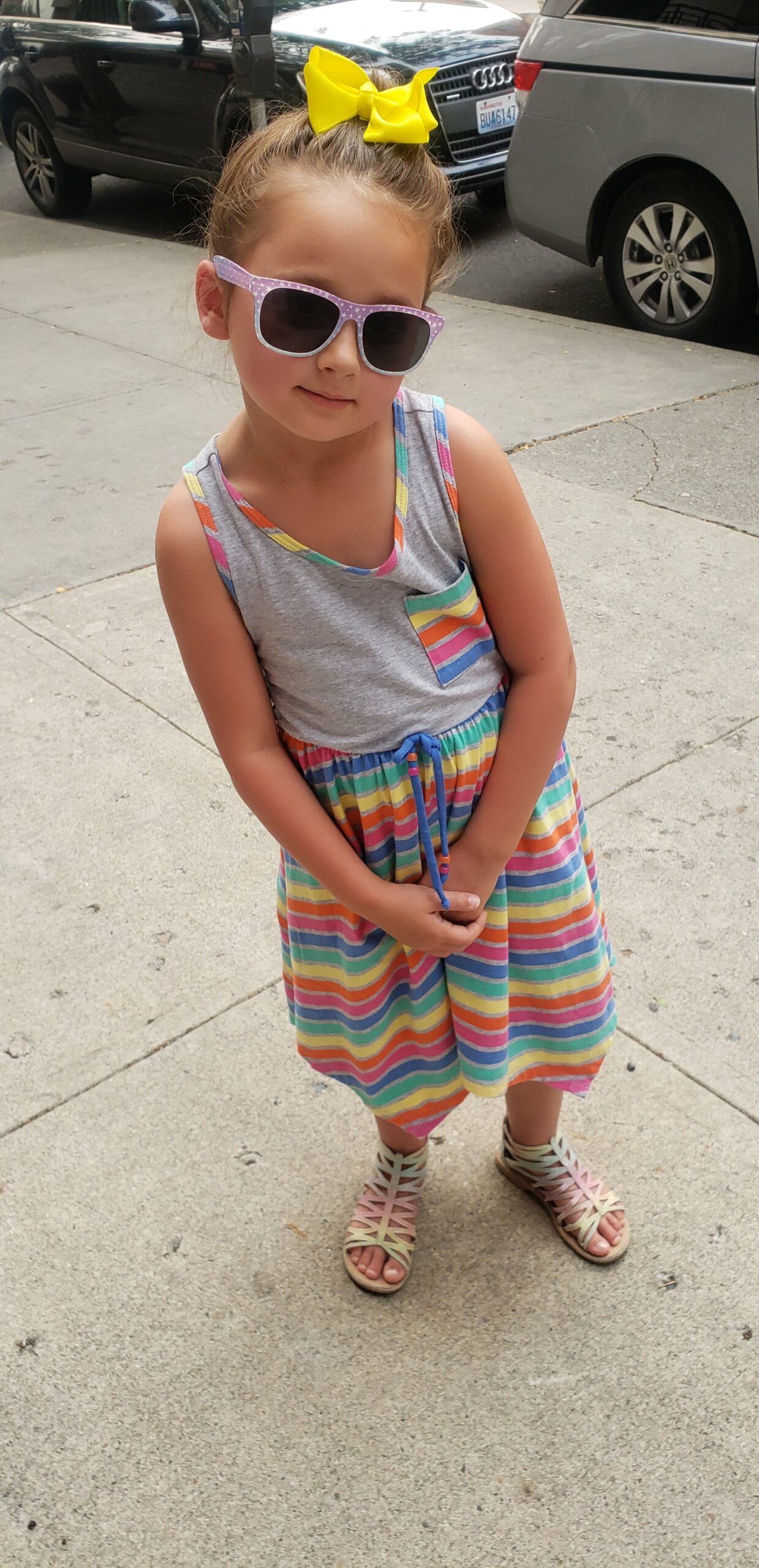 a young girl wearing sunglasses and a colorful striped dress stands confidently with her hands on her hips