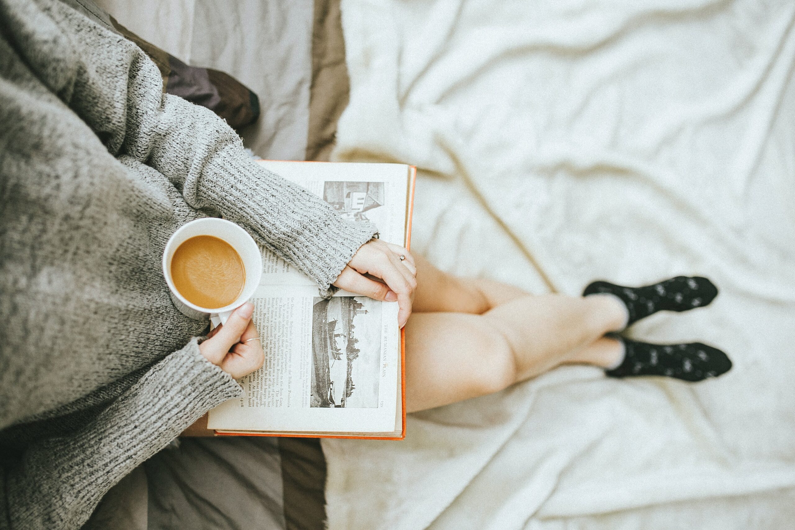 An image of a lady reading a book and drinking coffee while laying in her bed.